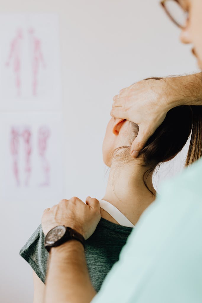 Back view of crop physician in uniform and wristwatch checking up neck of slim woman in doctor office.